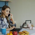 Woman working on a laptop at her kitchen table while on the phone